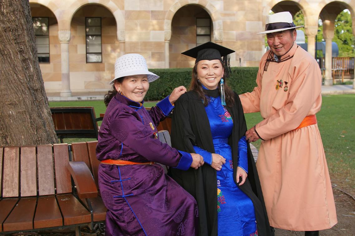 A Mongolian woman in a graduation cap and gown flanked by her parents who are wearing tradional Mongolian clothing.